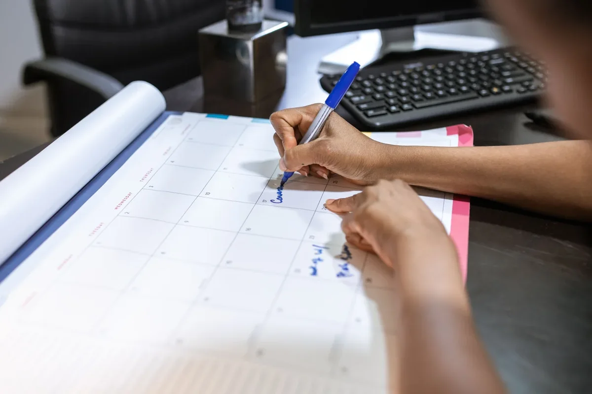 Person writing on a desk calendar beside a computer