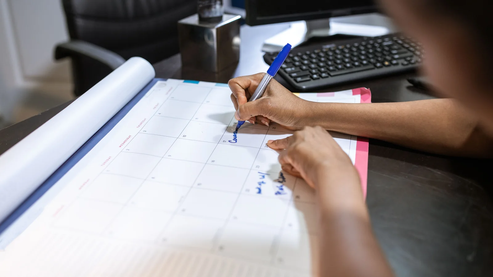 Person writing on a desk calendar beside a computer